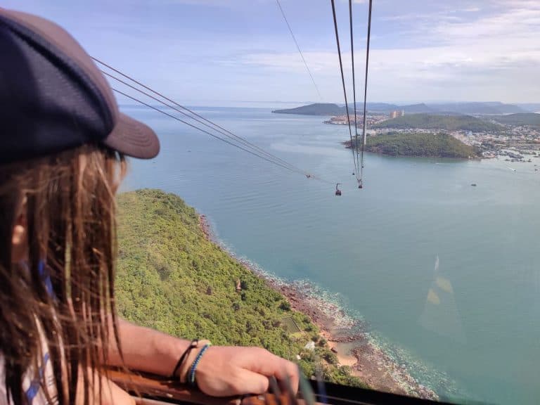 A child looks out from a cable car high above turquoise water, with long cable lines stretching toward distant islands and coastal towns. Green hills, rocky shoreline, and scattered boats fill the panoramic view below. This experience showcases one of the most scenic things to do in Phu Quoc Vietnam.