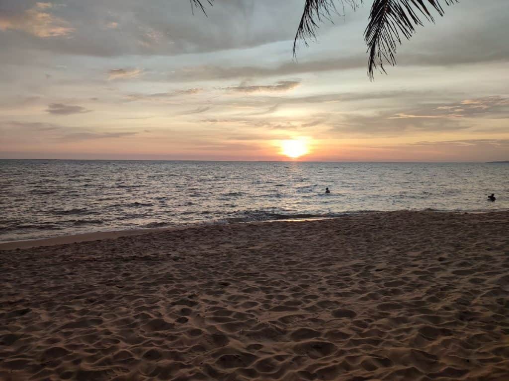 A long stretch of golden sand with calm waves on the shore and an orange sun setting in the distance in Phu Quoc, Vietnam