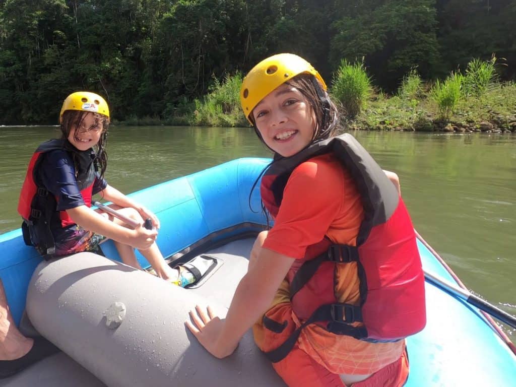 Two boys in life jackets and helmets sit in the front of a blue inflatable raft on the Sarapiqui River in Costa Rica