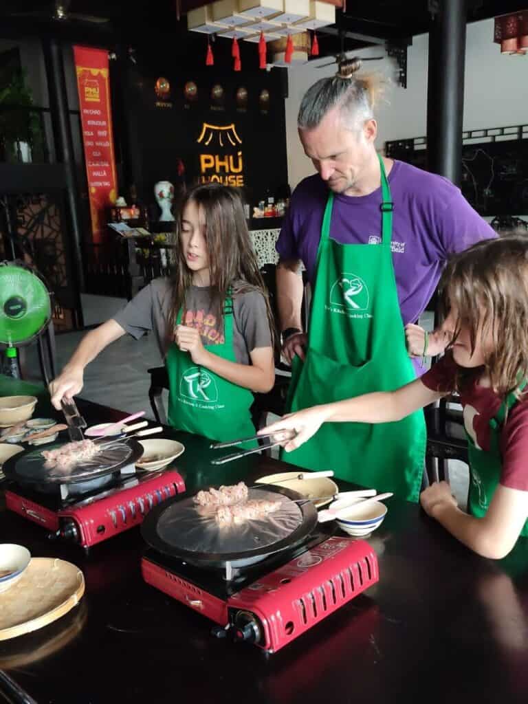 Two children and an adult wearing green aprons stand at a cooking station grilling meat on tabletop gas burners during a hands-on lesson. The family concentrates on turning the food while the instructor’s restaurant setting appears in the background. This scene shows a fun and interactive moment from a Phu Quoc cooking class.