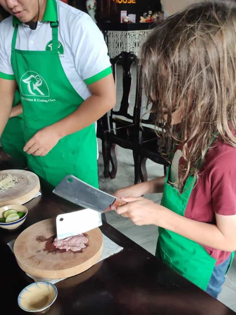 Young boy in green apron chops meat on a wooden board using two meat cleavers at a cooking class in Phu Quoc, Vietnam.
