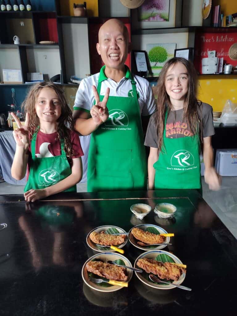 Chef and two young boys in green aprons stand behind a table smiling, with four dishes of Vietnamese food in front of them on the table at a Phu Quoc cooking class in Vietnam.