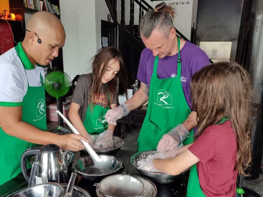 A cooking instructor guides an adult and two children as they mix ingredients in metal bowls, all wearing aprons and plastic gloves while working at a shared counter. The group leans in with focus as they stir and prepare a dish together. This image highlights the family-friendly experience of joining a Phu Quoc cooking class.