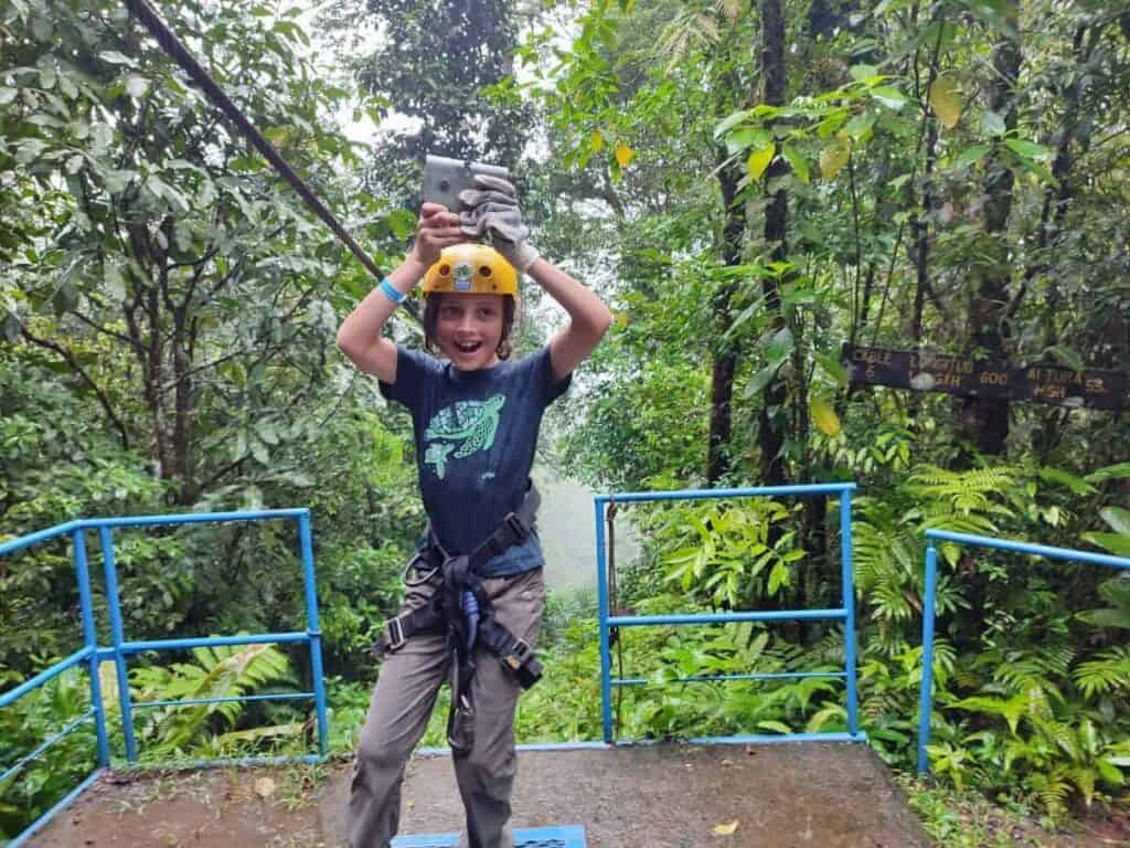 Excited child ready to zipline in La Fortuna, Costa Rica, wearing a helmet and harness at a rainforest canopy platform. Surrounded by lush greenery, the child smiles as they hold a phone, capturing the adventure. La Fortuna is renowned for its thrilling ziplining experiences, offering stunning views of the jungle and Arenal Volcano, making it a must-do family activity