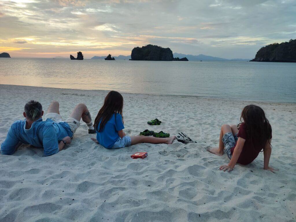A man and two young boys sit leaning back on a white sand beach in Langkawi, Malaysia. In the distance are several large rock formations in the ocean; the sun is setting behind them.