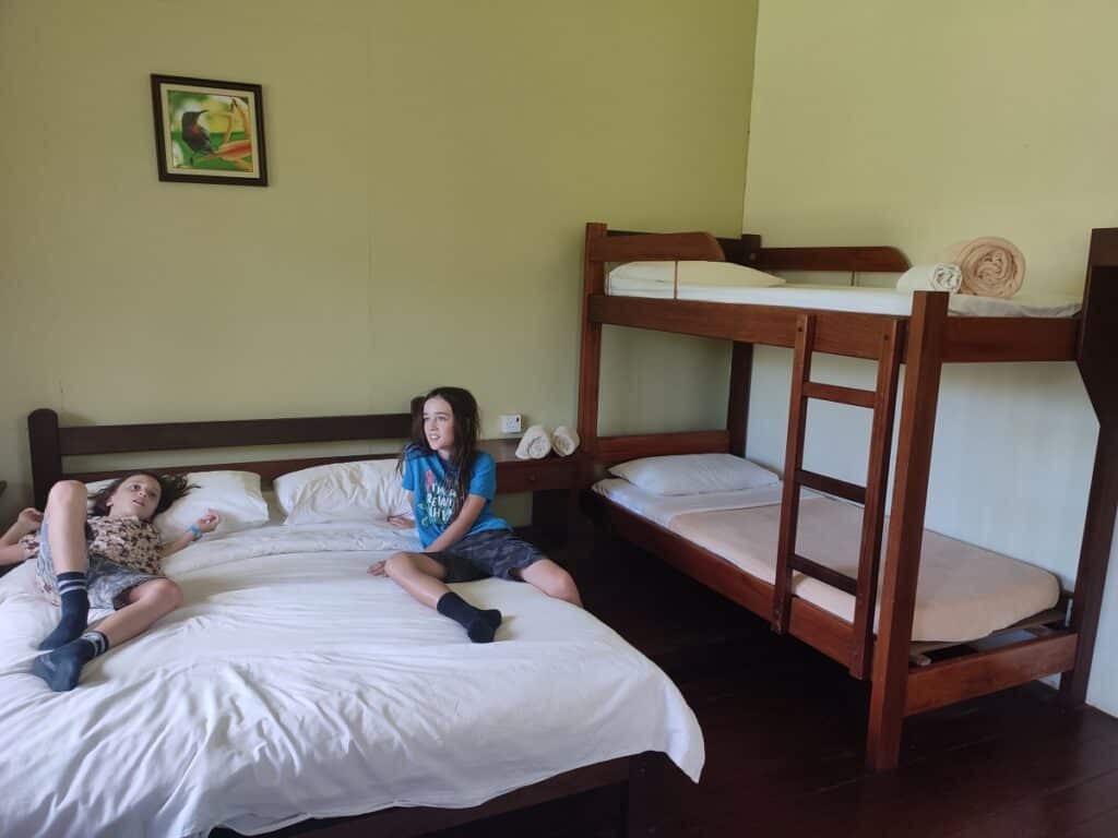 Two boys sit on a double bed chatting in a room at Sepilok B&B in Borneo. There is also a set of freshly made up bunk beds to the right of them, a polished wooden floor and a small picture on the wall above the main bed.