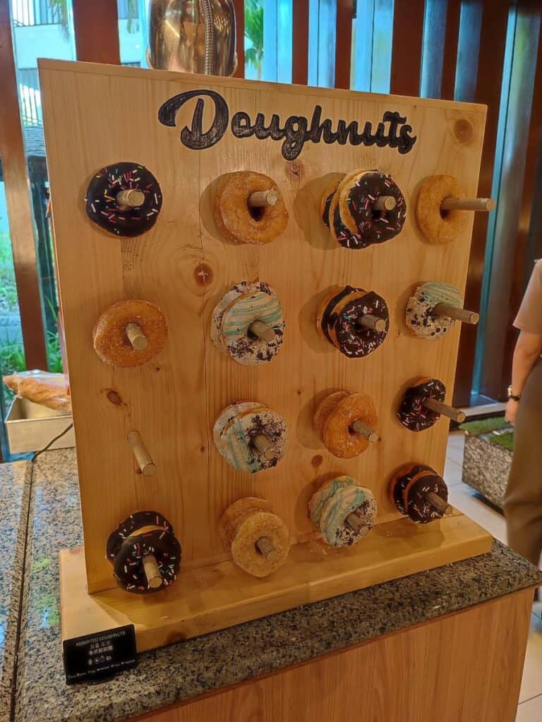 A wooden wall with the word 'Doughnuts' written across the top, with 16 small wooden sticks that a range of chocolate, iced and sugared ring doughnuts are hanging on. Part of the breakfast on offer at the Shangri-La Rasa Ria in Borneo.