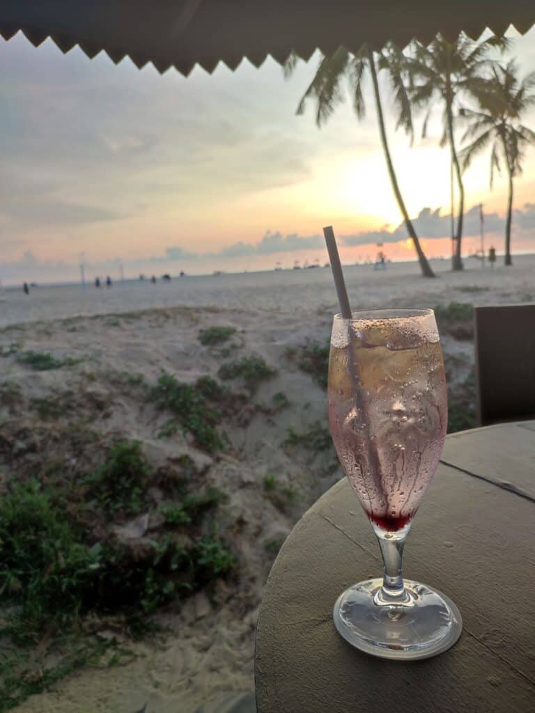 A long glass of pink cocktail with ice and a straw sits in the foreground, with the white sand beach and palm trees of the Shangri-La Rasa Rica beach in the background, The sun is setting casting pink and orange hues across the sky.
