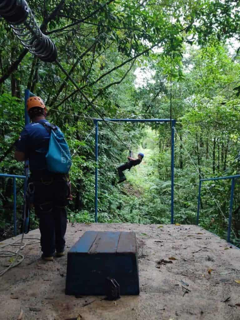 A person wearing a helmet zips across a line through the rainforest while a guide stands on the platform preparing the next rider. This action shot captures the excitement and safety setup that make this experience part of the best ziplining in La Fortuna.