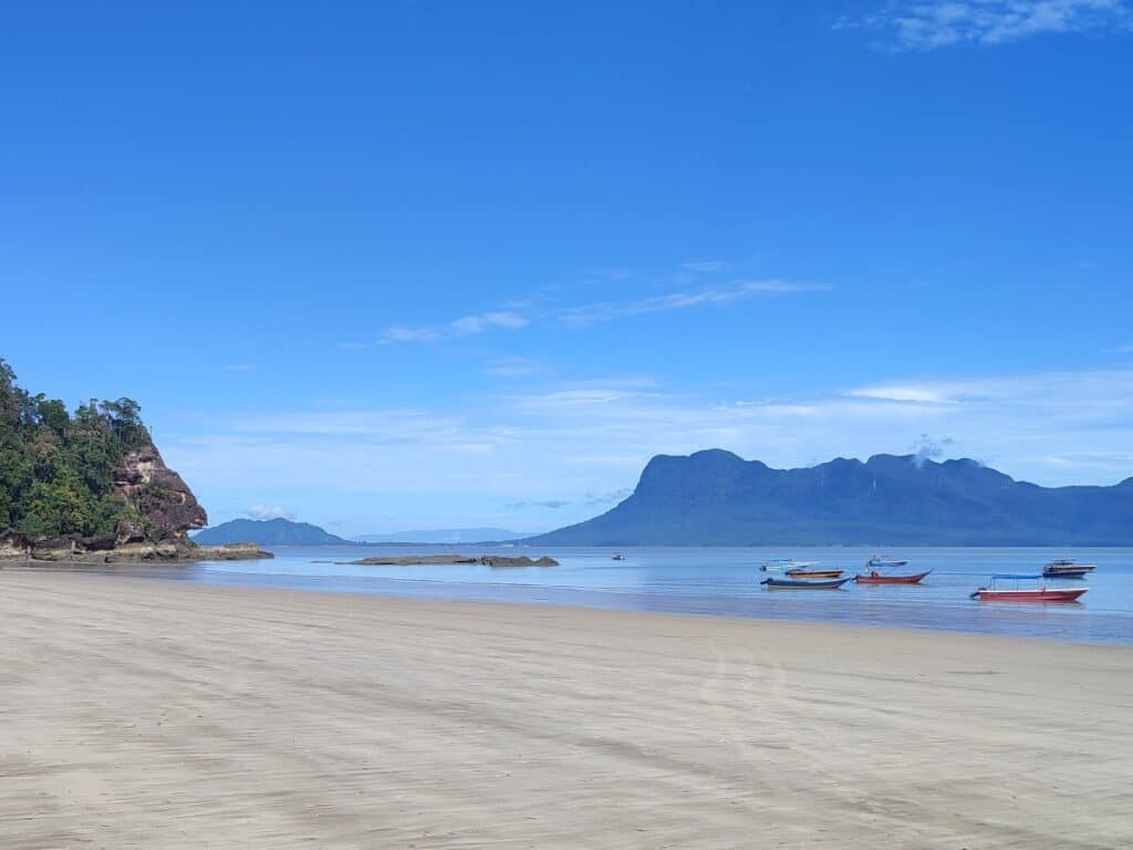 Wide sandy beach at Bako National Park with colorful wooden boats resting in shallow water, rugged cliffs on the left, and misty mountains across the sea under a bright blue sky. The calm shoreline near Kuching shows the scenic coastal landscape families can expect when visiting Bako National Park in Borneo.