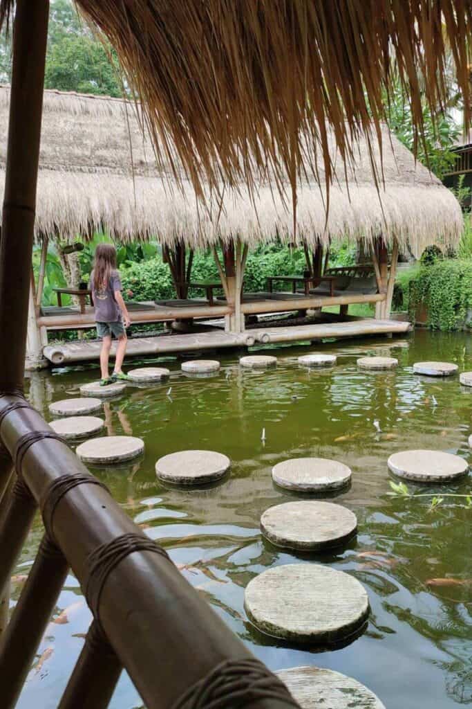 A child steps across round stone platforms over a koi pond toward a thatched bamboo pavilion restaurant in Ubud. The tranquil water garden atmosphere showcases a unique and scenic option for where to eat in Ubud.