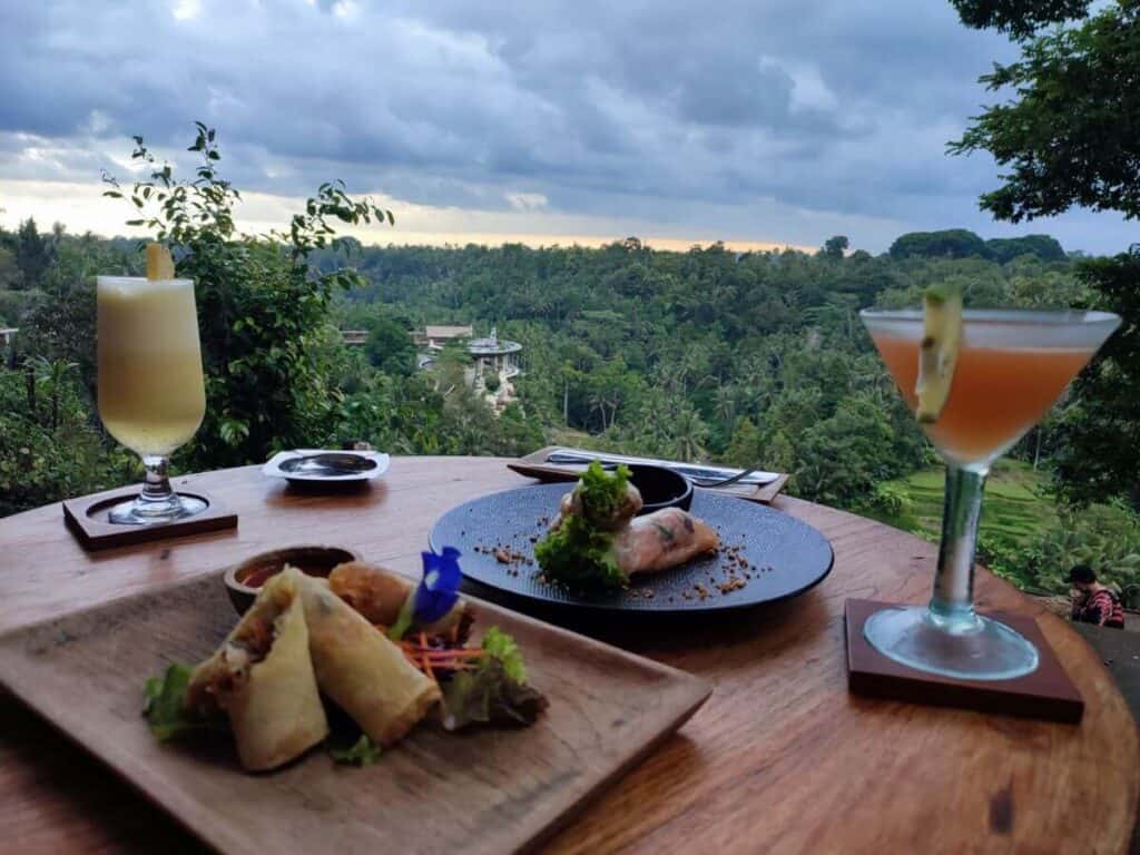 Plates of spring rolls and cocktails sit on a wooden table overlooking lush jungle valley views in Ubud at sunset. This scenic dining setting captures the kind of unforgettable experience travelers look for when deciding where to eat in Ubud.