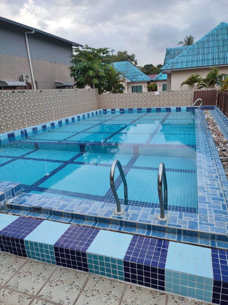 Rectangular outdoor swimming pool at D'Villa Guesthouse in Langkawi, Malaysia. Small trees are dotted around the edge, and low-rise buildings with green roofs are in the background.