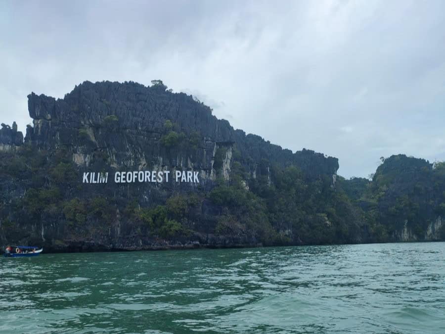 A huge limestone karts formation rises from the aquamarine sea near Langkawi, Malaysia. White lettering on the side of the rock states 'Kilim Geoforest Park. The rest of the rock is covered in green vegetation. Viewed from a boat on a Langkawi mangrove tour.