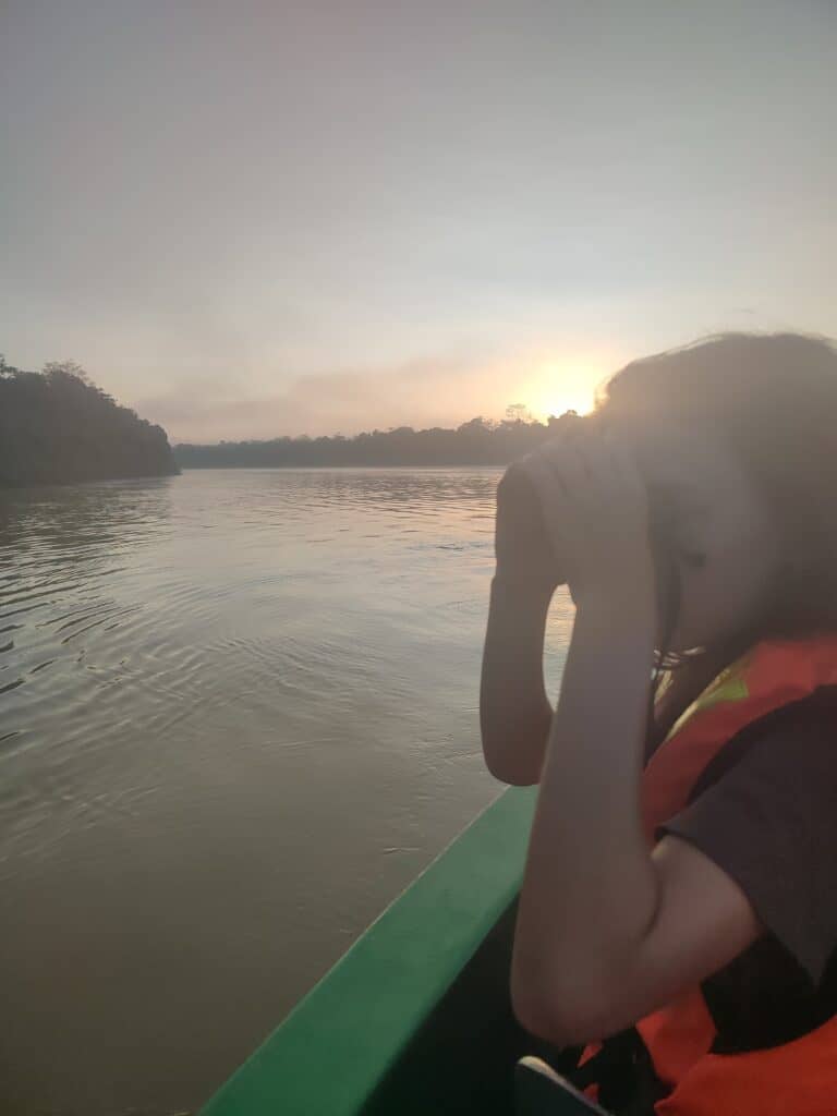 A boy in a life jacket sits at the front of a small boat looking for wildlife through binoculars at sunrise over the Kinabatangan River. Calm water reflects the early morning light during a Kinabatangan River tour.