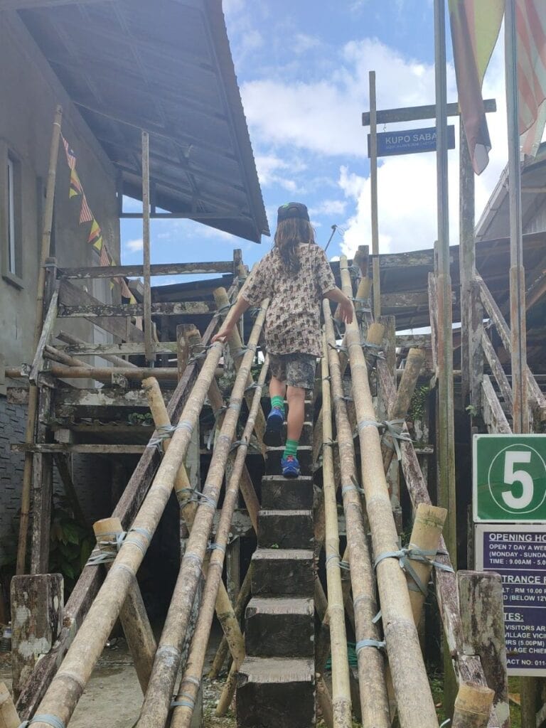 Child walking up a narrow bamboo staircase between rustic wooden buildings in a traditional Bidayuh village setting outside Kuching Borneo. The scene captures a cultural stop on a Kuching itinerary with weathered wood textures and bright blue sky overhead.