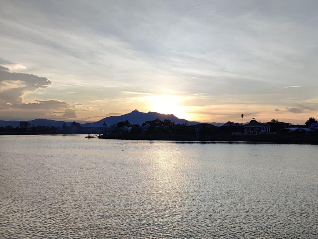 Orange sunset across the wide river in Kuching, Borneo. In the distance are the peaks of mountains, with several low-rise buildings and palm trees on the far shore of the water.