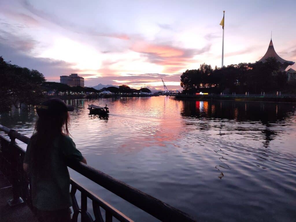 Silhouette of a child looking out over the Sarawak River at sunset in Kuching Borneo with pink and orange skies reflecting on the water and a small boat drifting past. The peaceful waterfront view showcases a scenic moment to include in a Kuching itinerary.