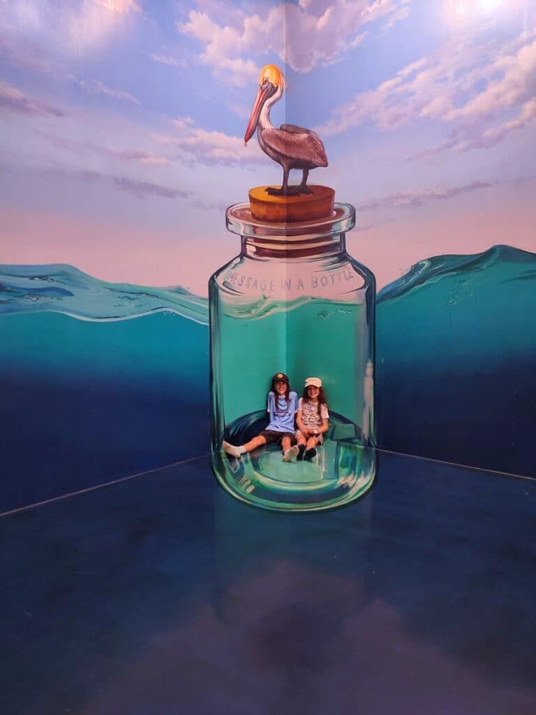 Two children sitting inside a large painted message in a bottle mural with a pelican perched on top and ocean waves surrounding the scene at a 3D art museum in Langkawi. This playful interactive art spot adds a fun photo opportunity to a Langkawi Itinerary for families.