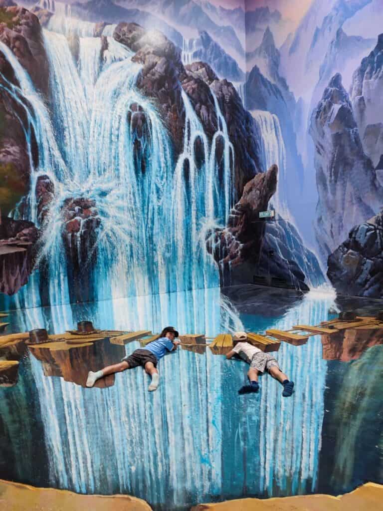 Two children lying across a realistic waterfall illusion mural that makes it look like they are hanging over cascading blue water and rocky cliffs in Langkawi. This immersive 3D art experience is a creative indoor activity to include in a Langkawi Itinerary for families.