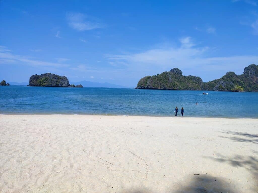 Wide stretch of soft white sand at Tanjung Rhu Beach with two children walking toward calm turquoise water and small forested islands rising in the distance under a bright blue sky. This peaceful beach in Langkawi is perfect for swimming and exploring with kids on a Langkawi itinerary for families.