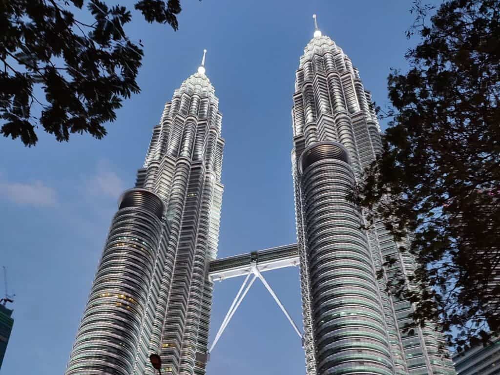 The Petronas Twin Towers rise into the evening sky, connected by the skybridge and framed by nearby trees. Visiting this iconic landmark at ground level is a popular and free activity when exploring Kuala Lumpur on a budget.