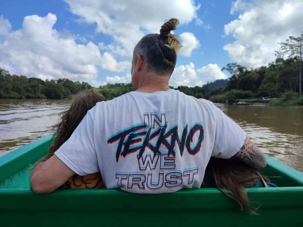 Man in white t shirt sits at the front of a small boat on the muddy brown Kinabatangan River in Borneo, with his arms around two children, one on each side of him.