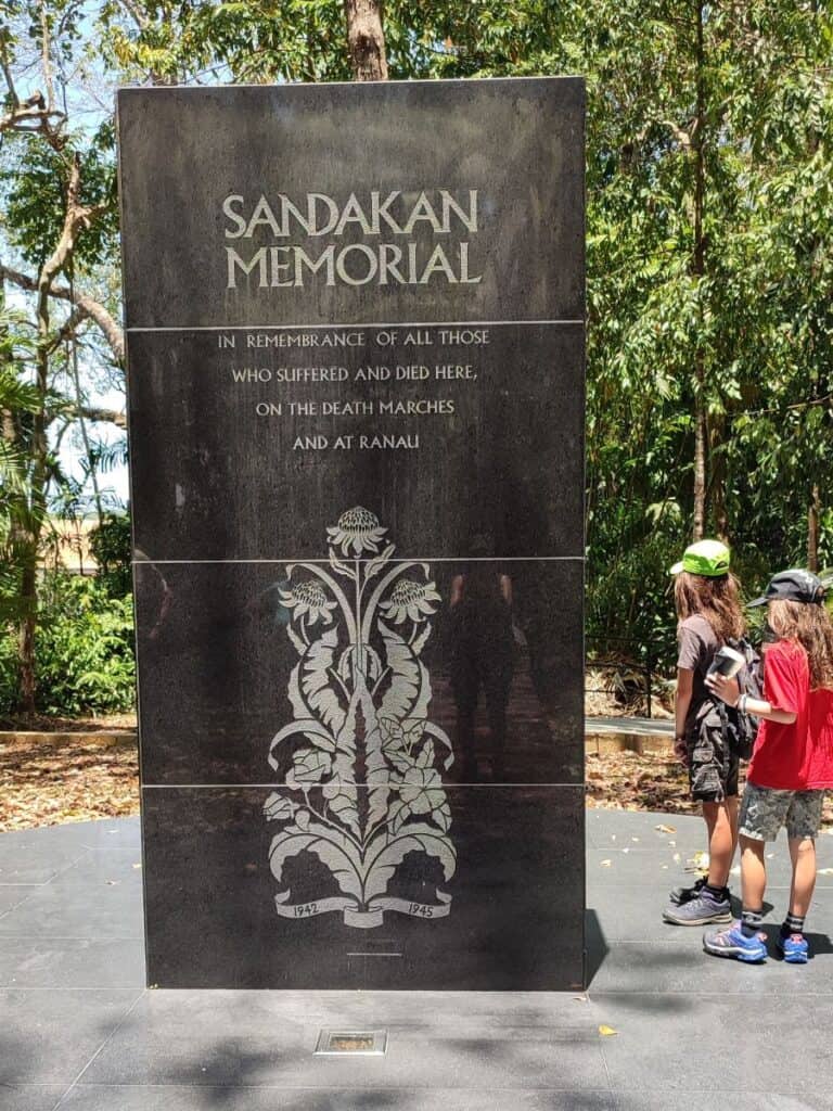 Two children stand to the right of the rectangular Sandakan Memorial plaque that commemorates the site of the WWII Prisoner of War Camp in Sandakan, Borneo and those who lost their lives there.