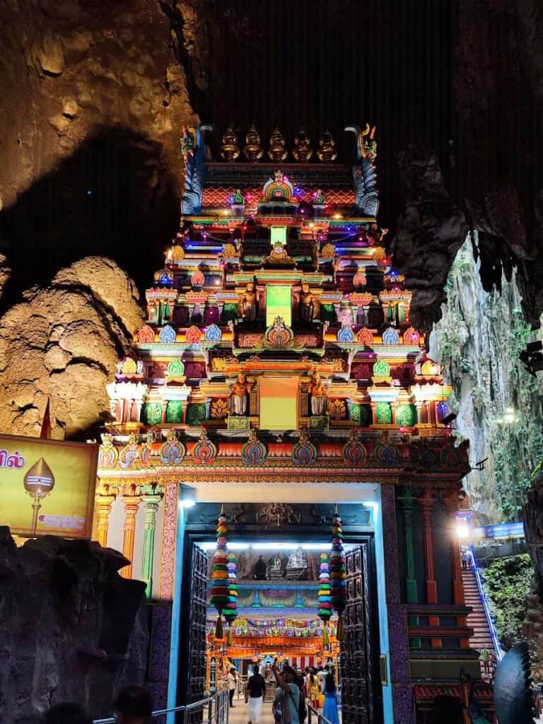 Colorful Hindu temple entrance built into the limestone cave at Batu Caves, covered in bright carvings and lights as visitors walk inside beneath the towering structure. The ornate gateway highlights the dramatic setting of visiting Batu Caves where the temple sits directly within the cavern walls.