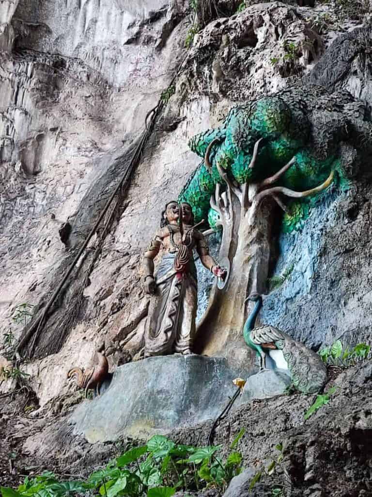 Hindu statue scene set against the limestone walls inside Batu Caves featuring a figure standing beside a sculpted tree with a peacock and rooster at the base. Religious artwork carved into the cave environment that visitors encounter when visiting Batu Caves.