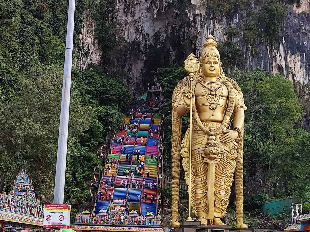 The towering golden statue of Lord Murugan stands at the entrance to Batu Caves with the famous rainbow colored staircase rising behind it into the limestone cliff. Visitors climb the vibrant steps during their experience visiting Batu Caves in Kuala Lumpur.