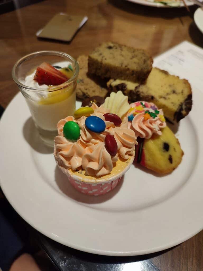 Plate of desserts at Shangri La Kuala Lumpur including a frosted cupcake topped with colorful chocolate candies slices of marble cake a small glass of creamy pudding with strawberry and a decorated mini sponge cake. Close up view of the sweet treats served at the Horizon Lounge, with access included in a Family Suite stay.