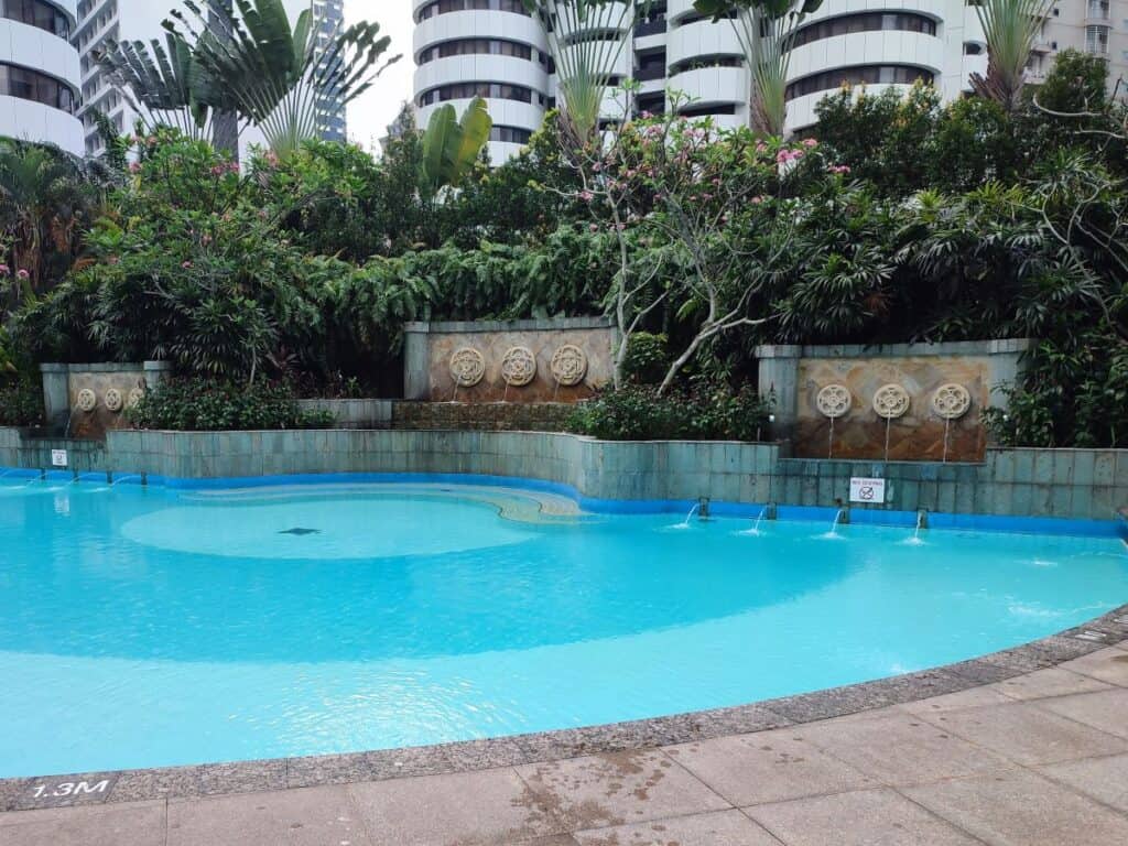 Outdoor swimming pool at Shangri La Kuala Lumpur surrounded by lush tropical plants and modern high rise buildings. Curved turquoise pool with small water fountains along a stone wall creating a relaxing hotel atmosphere.