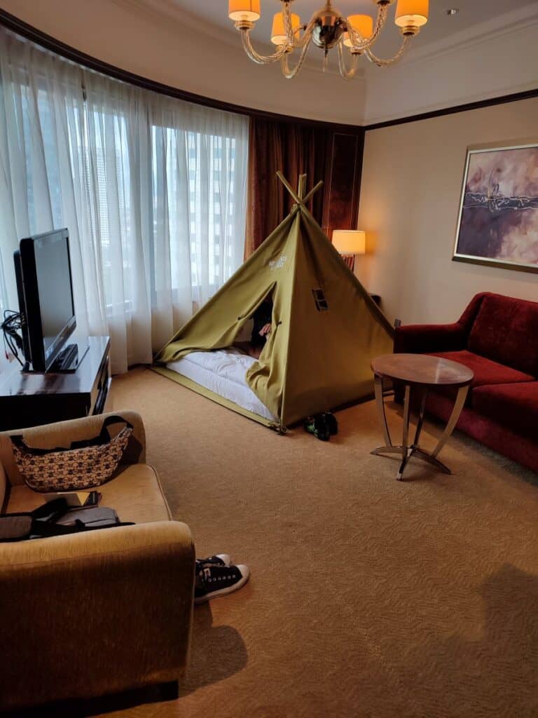 Spacious hotel room at Shangri La Kuala Lumpur with a small indoor camping style tent set up on the floor beside the bed for a child. Living area includes a sofa armchair chandelier and large windows letting in daylight as part of a family stay featured in this Shangri La Kuala Lumpur review.