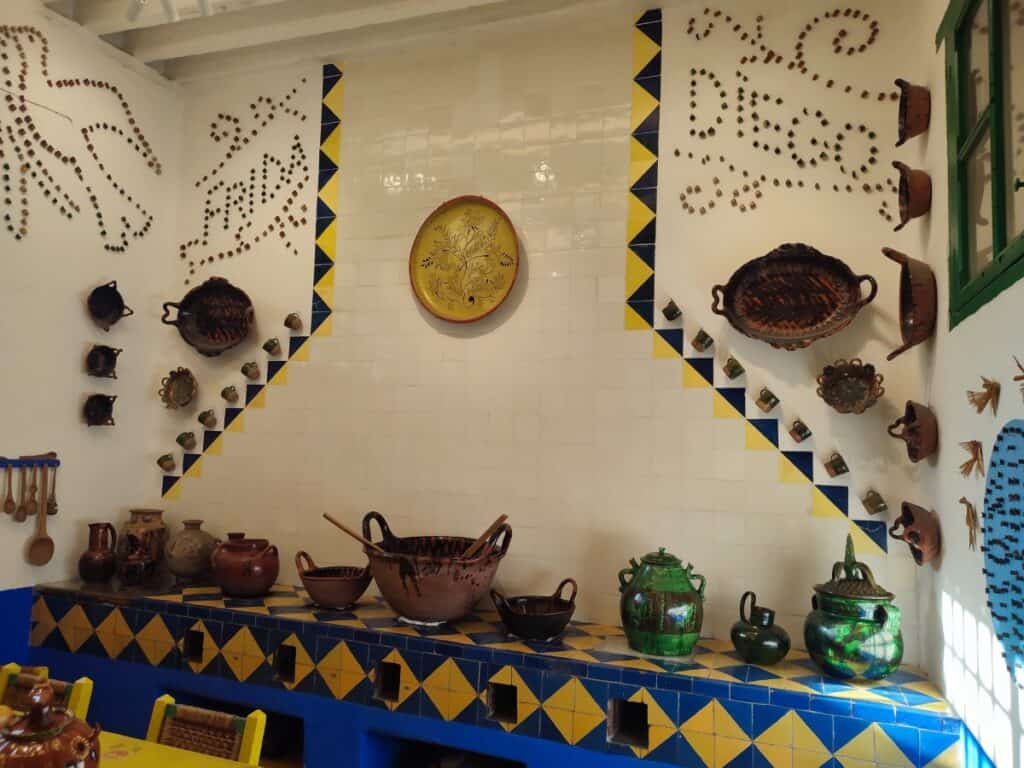 The vibrant kitchen at La Casa Azul (the Frida Kahlo Museum) in Coyoacán, Mexico City. Blue and yellow diamond-patterned tiles cover the countertop and walls, which are decorated with traditional clay pots, bowls, and cookware. The names 'Frida' and 'Diego' are spelled out in small clay figures pinned directly to the white tiled wall above.