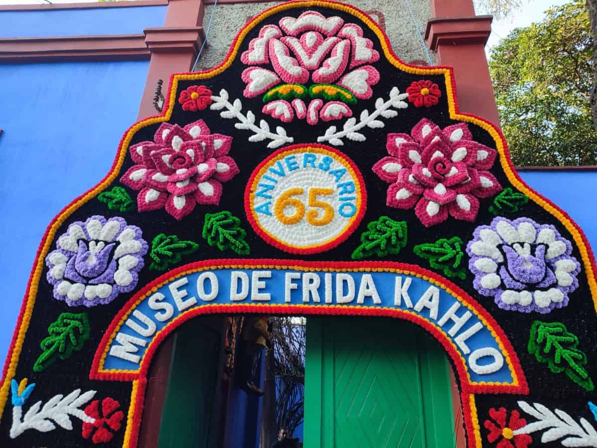 Colorful floral arch at the entrance of the Museo de Frida Kahlo with bright pink, white, and red flowers surrounding a circular sign that reads “ANIVERSARIO 65” and “MUSEO DE FRIDA KAHLO,” highlighting a cultural landmark often included in things to do in Mexico City.