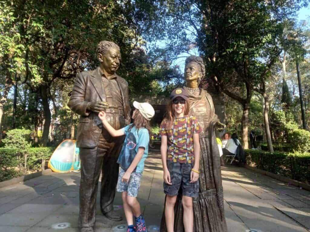 Two children pose between bronze statues of Diego Rivera and Frida Kahlo in Jardín Centenario, Coyoacán, Mexico City. One child touches Rivera's hand while the other smiles beside Kahlo, surrounded by lush green trees