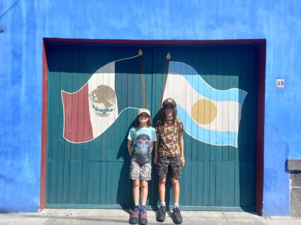 Two children standing in front of a large painted wooden door featuring the Mexican flag and another flag design, set against a vivid blue wall, capturing a casual street photo moment during things to do in Mexico City.