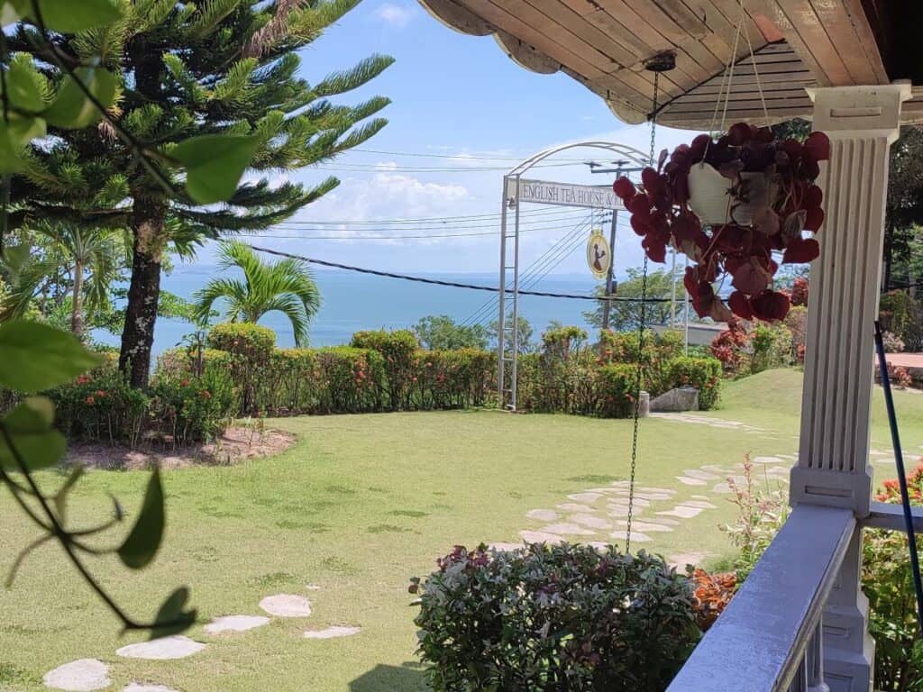 The lush hilltop garden of the English Tea House and Restaurant in Sandakan, Borneo, with a stone pathway, tropical flowers, and a sweeping view of the Sulu Sea visible beyond the grounds — a peaceful stop on any Sandakan itinerary