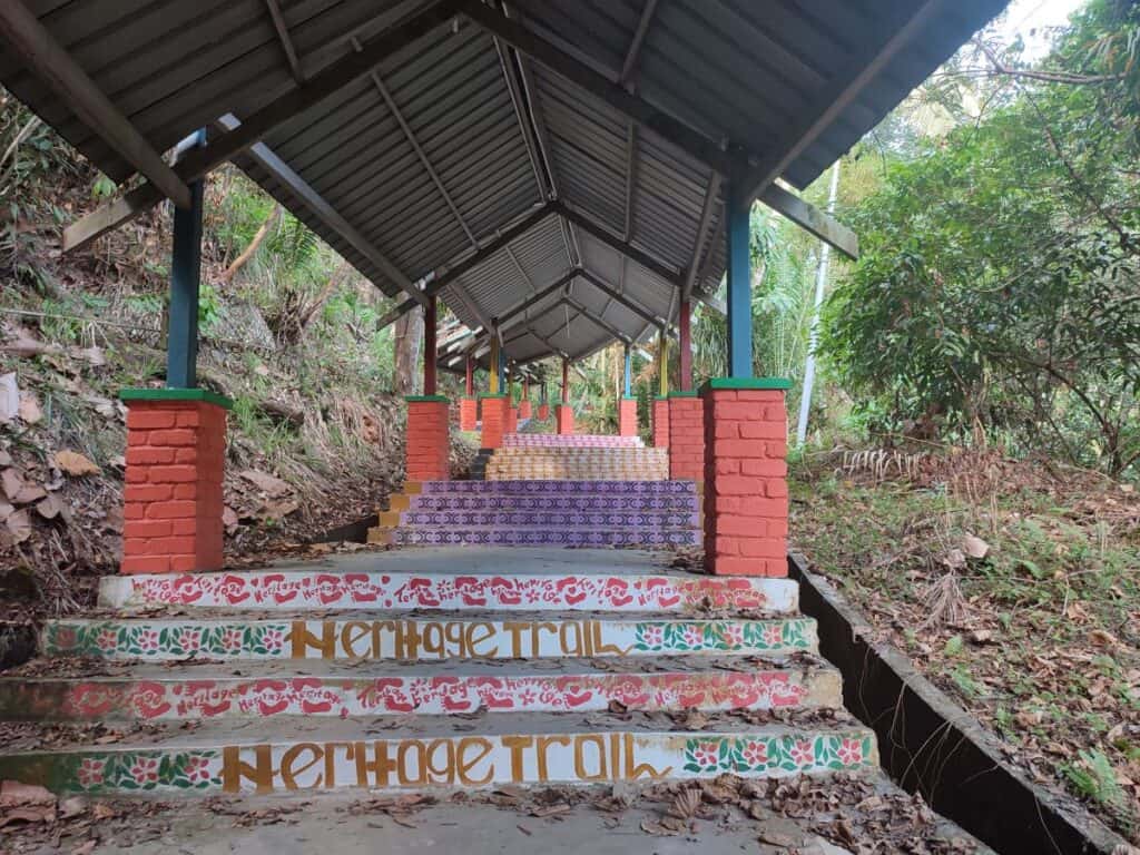 Covered staircase on the Sandakan Heritage Trail in Borneo, with steps decorated in colourful painted tiles and the words 'Heritage Trail' written along the risers, surrounded by tropical jungle vegetation.