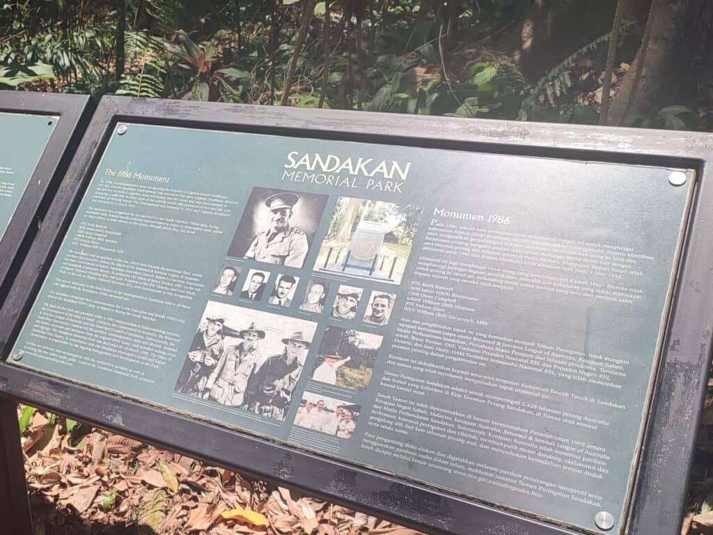 An information board at Sandakan Memorial Park in Borneo, titled 'The 1986 Monument,' with bilingual English and Malay text and historical photographs commemorating the Australian and British POWs who endured the Sandakan Death Marches during World War II.