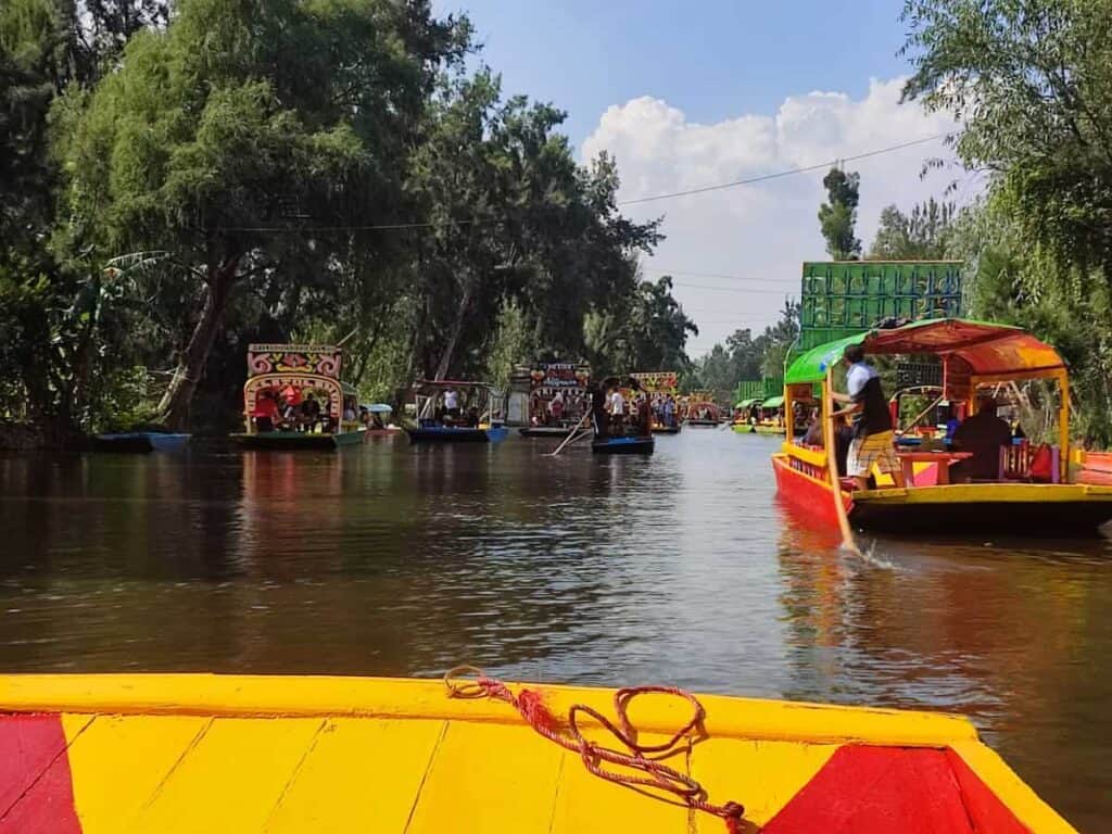 View from the prow of a traditional wooden boat - in yellow and red - on the Xochimilco canals in Mexico City. In the distance a number of other yellow and red boats drift on the muddy canal waters, with green leafy trees lining the shoreline on either side.