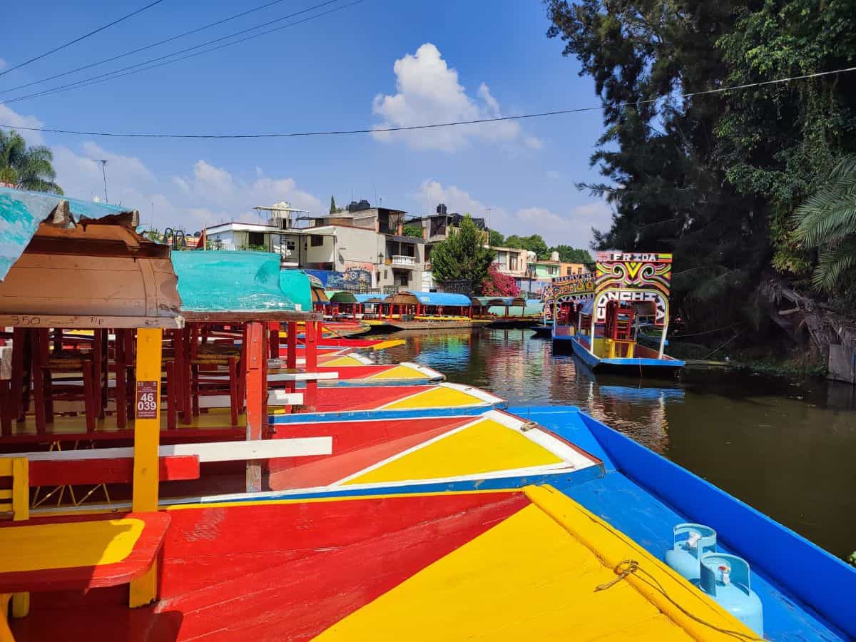 Brightly painted trajinera boats in red, yellow, and blue are moored side by side along the Xochimilco canals in Mexico City. One boat is named 'Frida.' Low-rise buildings, trees, and more colorful boats line the far bank of the calm, narrow waterway.