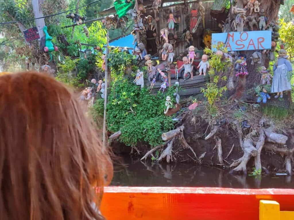 Seen from the deck of a trajinera boat on the Xochimilco canals, the famous Isla de las Muñecas (Island of the Dolls) comes into view — dozens of weathered, eerie dolls and doll parts hanging from trees and fencing along the muddy bank. A child with red hair watches from the foreground. Signs reading 'No Tirar Basura' and 'No Pasar' are visible among the dolls.