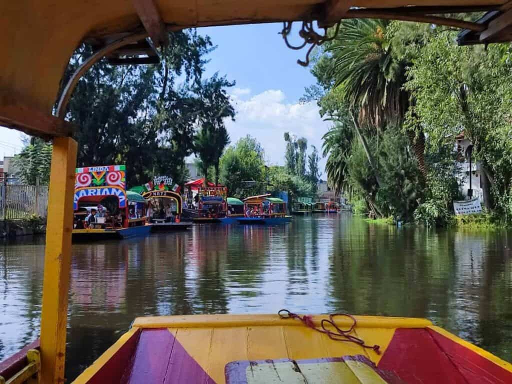 The view from the bow of a yellow and red trajinera boat cruising the tree-lined Xochimilco canals in Mexico City. Several other brightly painted and occupied trajineras are visible ahead on the calm waterway, with a gondolier poling a red and green boat on the right.