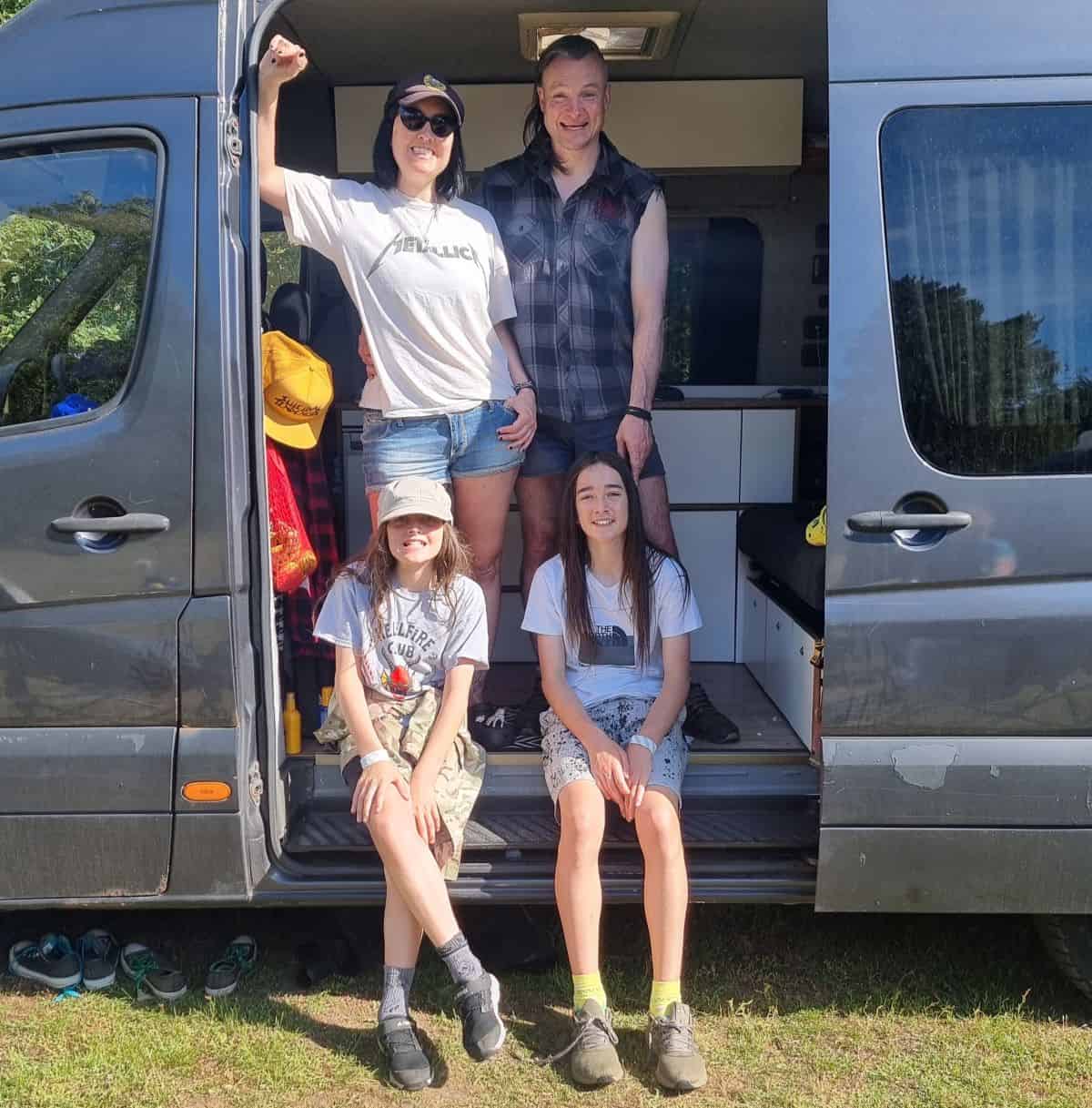Family of four - mum, dad and two boys - stand/sit in the open door of a grey Mercedes Sprinter campervan.