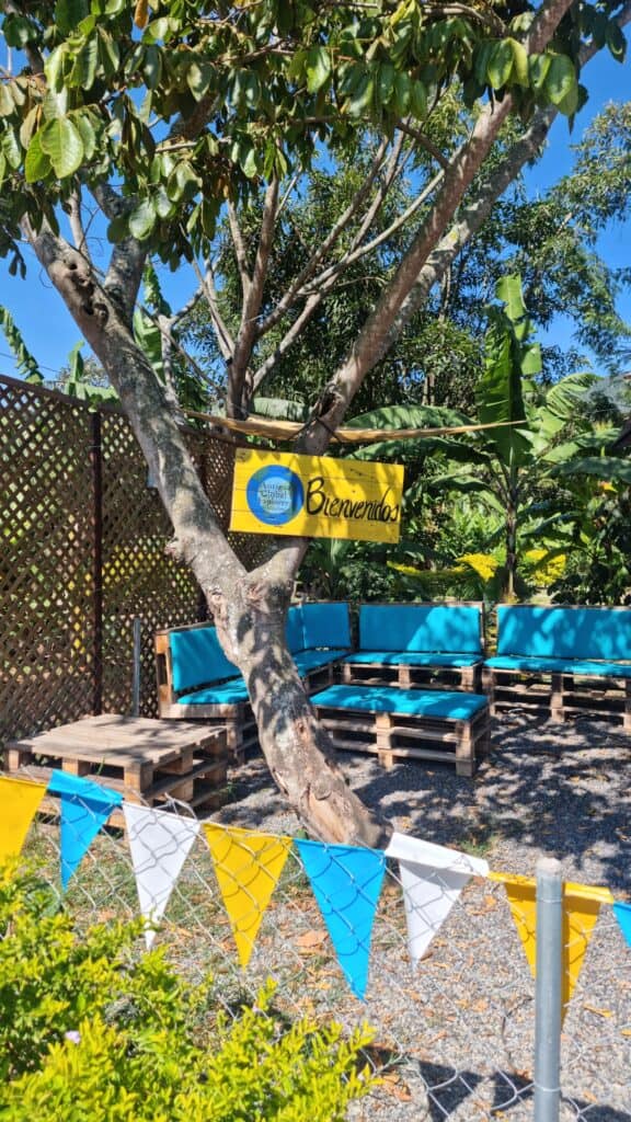 Tree with a yellow wooden sign fixed to it that says 'Bienvenidos' and identifies the location as Antigua Guatemala Explorers, a worldschooling hub in Guatemala. In the foreground is yellow and blue bunting along a fence and there is a small covered structure behind for children to do activities in.