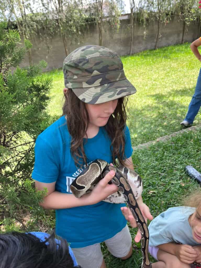 10-year-old boy in blue t shirt and camo baseball cap sits on the ground holding a large python.