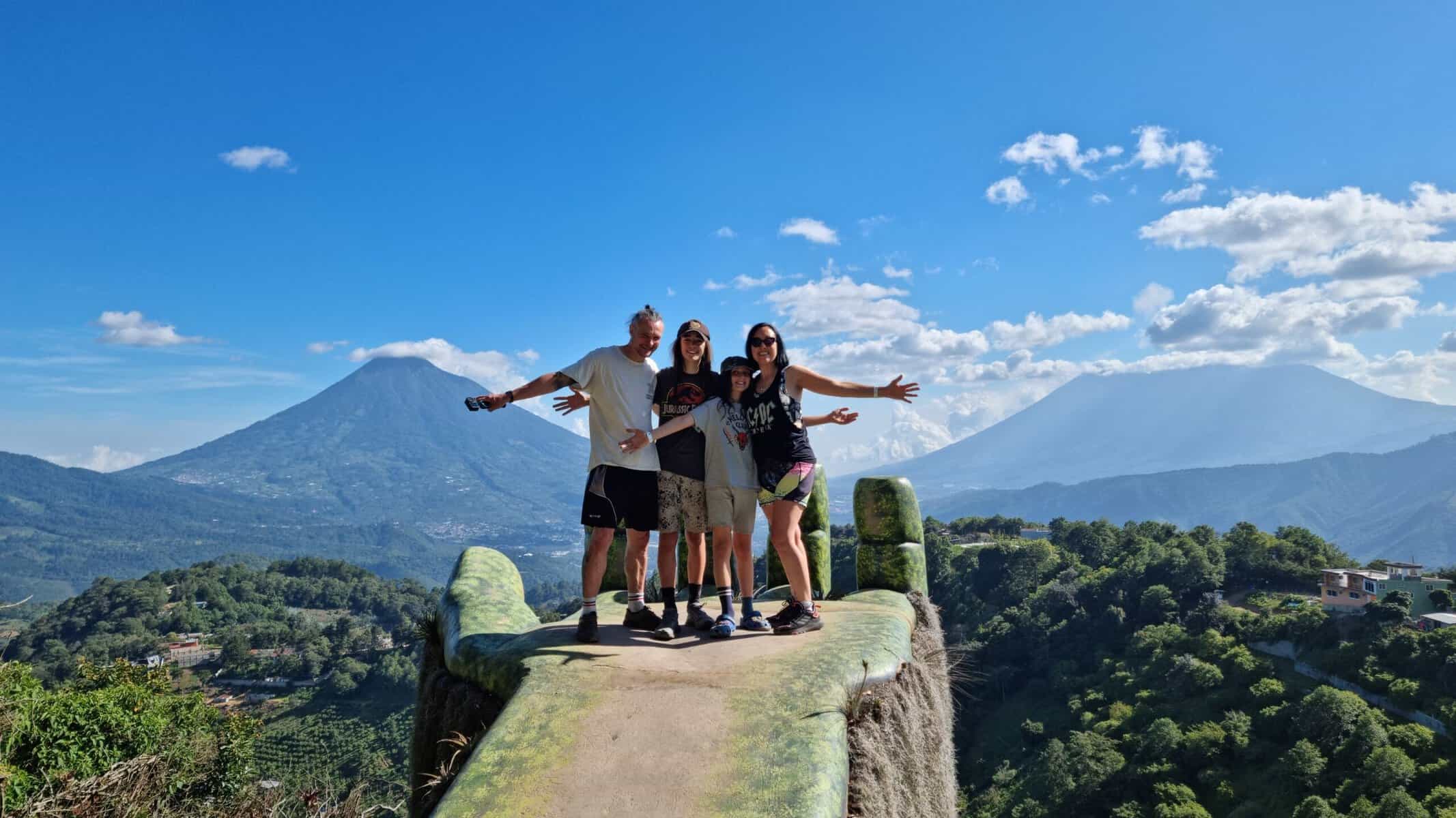 Family of four - man, woman and two boys - stand with their arms out-stretched inside a giant hand sculpture on a family gap year in Guatemala. In the background are two volcanoes and a densely forested landscape.