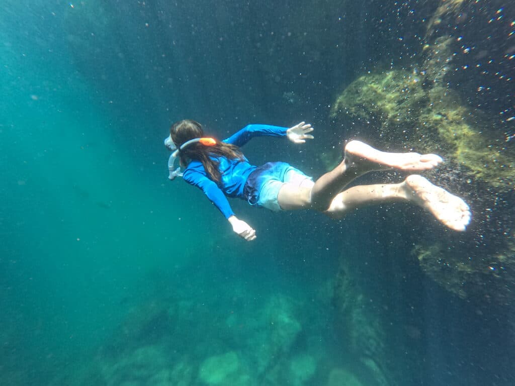 Child in blue rash vest and swim shorts swims in the green underwater at Las Grietas in Galapagos, one of our fave destinations for part of a family gap year.